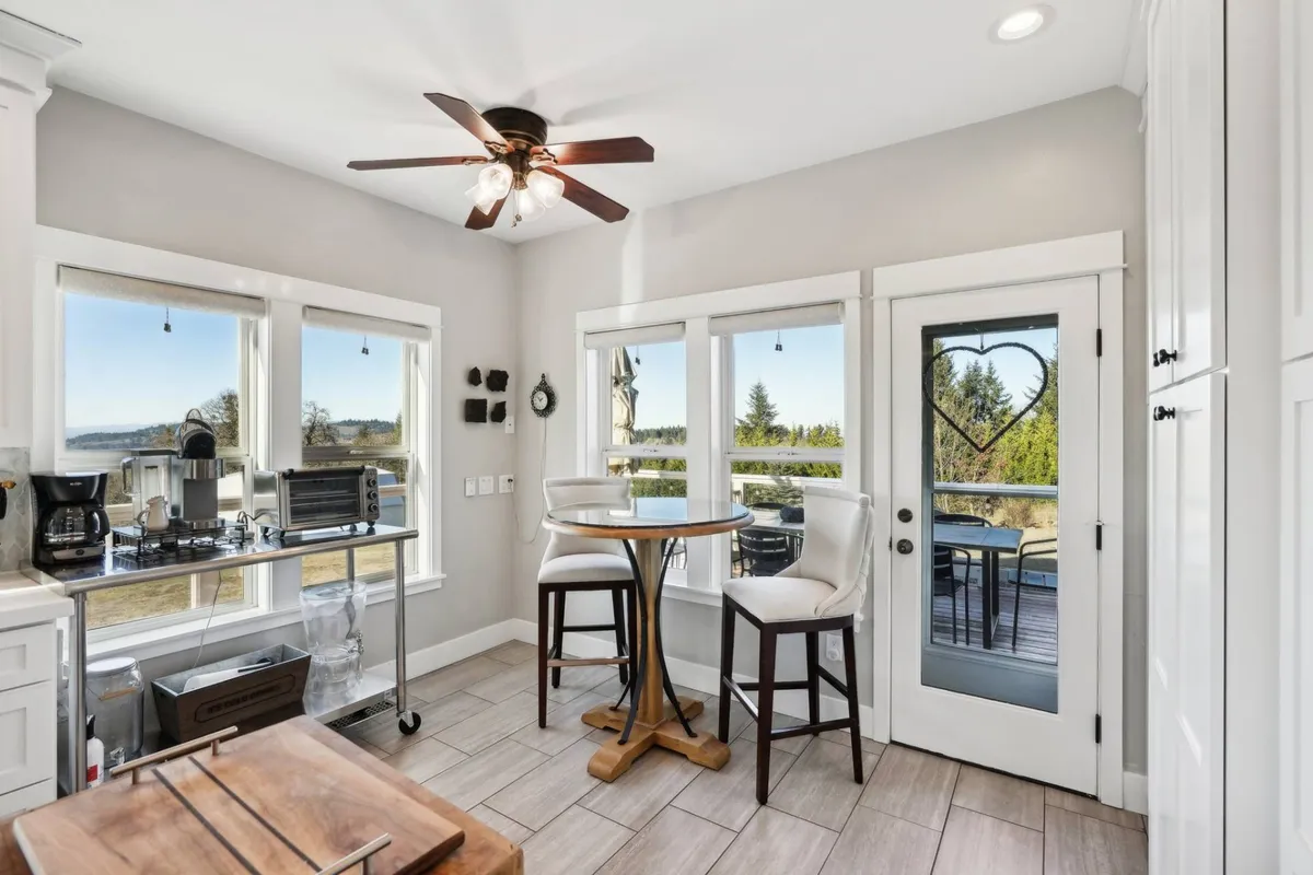 A bright kitchen area with a kitchen table positioned near large windows. The windows offer a view outside, filling the space with sunlight and connecting the inside with the outdoors.