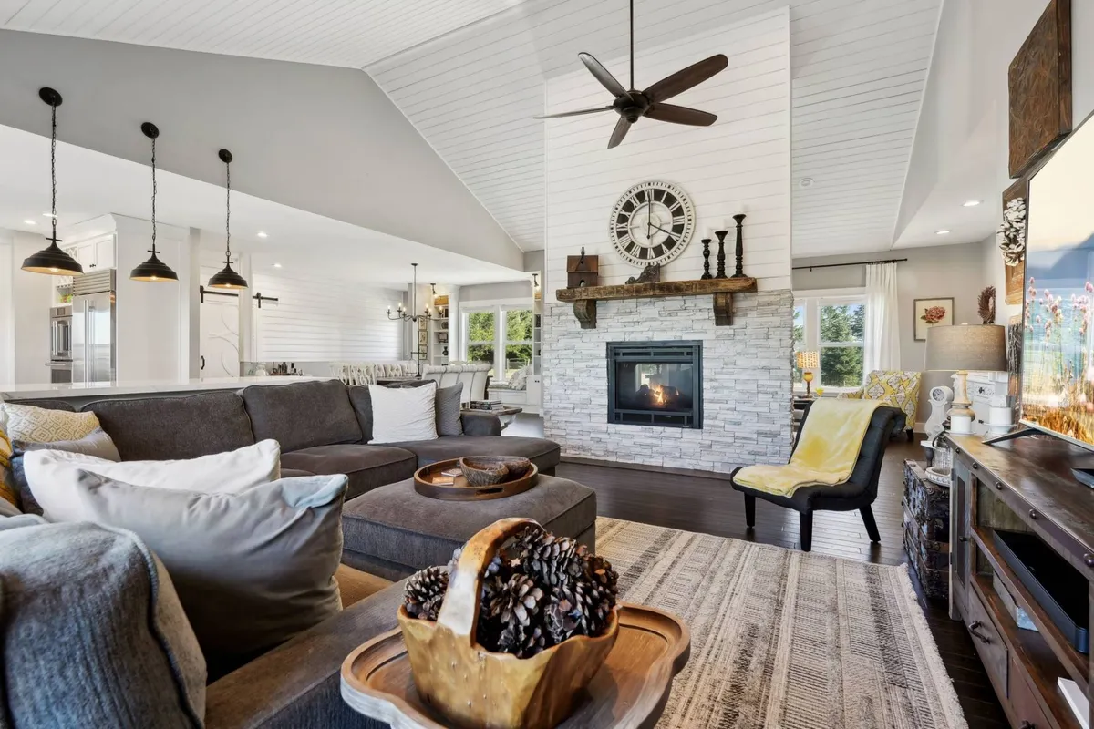 The image showcases a spacious, sunlit living room centered around a striking fireplace, with a plush, oversized sectional sofa inviting guests to gather and unwind. Decorative touches like a large clock and artfully arranged mantle décor add warmth and personality to the space, creating a perfect setting for cozy evenings, lively celebrations, or tranquil moments by the fire.