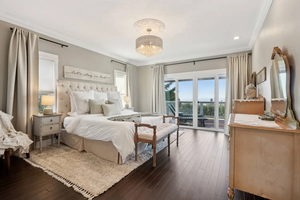 A serene bedroom featuring a plush bed positioned to enjoy soft natural light streaming in through large windows. Elegant touches like a chandelier and a dresser enhance the tranquil atmosphere, and the view through the windows offers a peaceful backdrop.