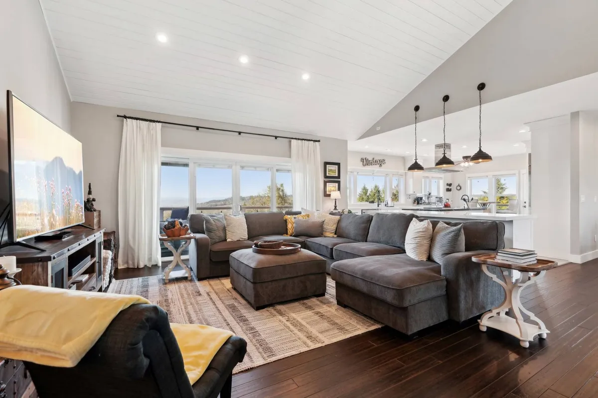 A spacious living area showing a large sectional sofa, a TV, and a dining area in the background, all surrounded by oversized windows that frame sweeping vineyard and mountain views. The furniture is plush and arranged for conversation and relaxation, creating a seamless flow between living, dining, and kitchen spaces.