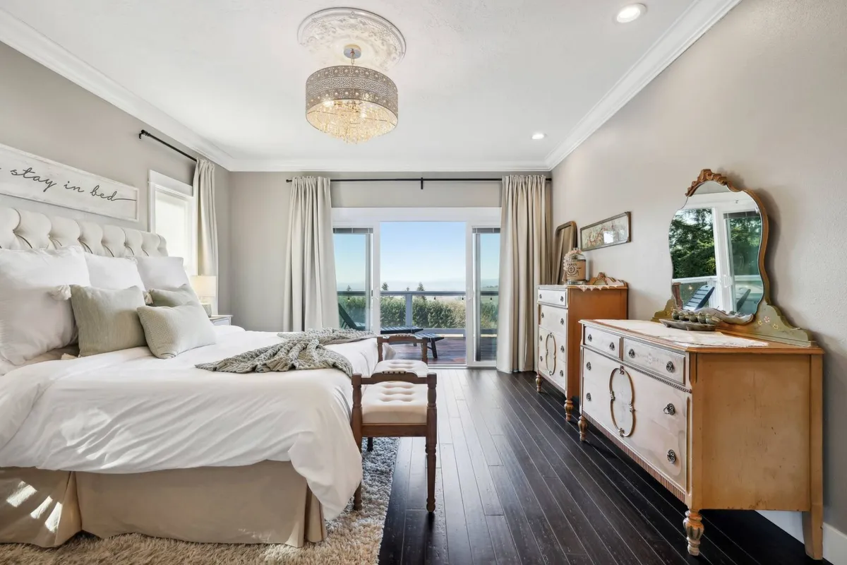 A calming bedroom with plush bedding, a chandelier, a small dressing table, and sliding doors that frame a sweeping vista of the outdoors. Natural light pours in, emphasizing the peacefulness of the space and inviting guests to enjoy a morning coffee while watching the sun rise.