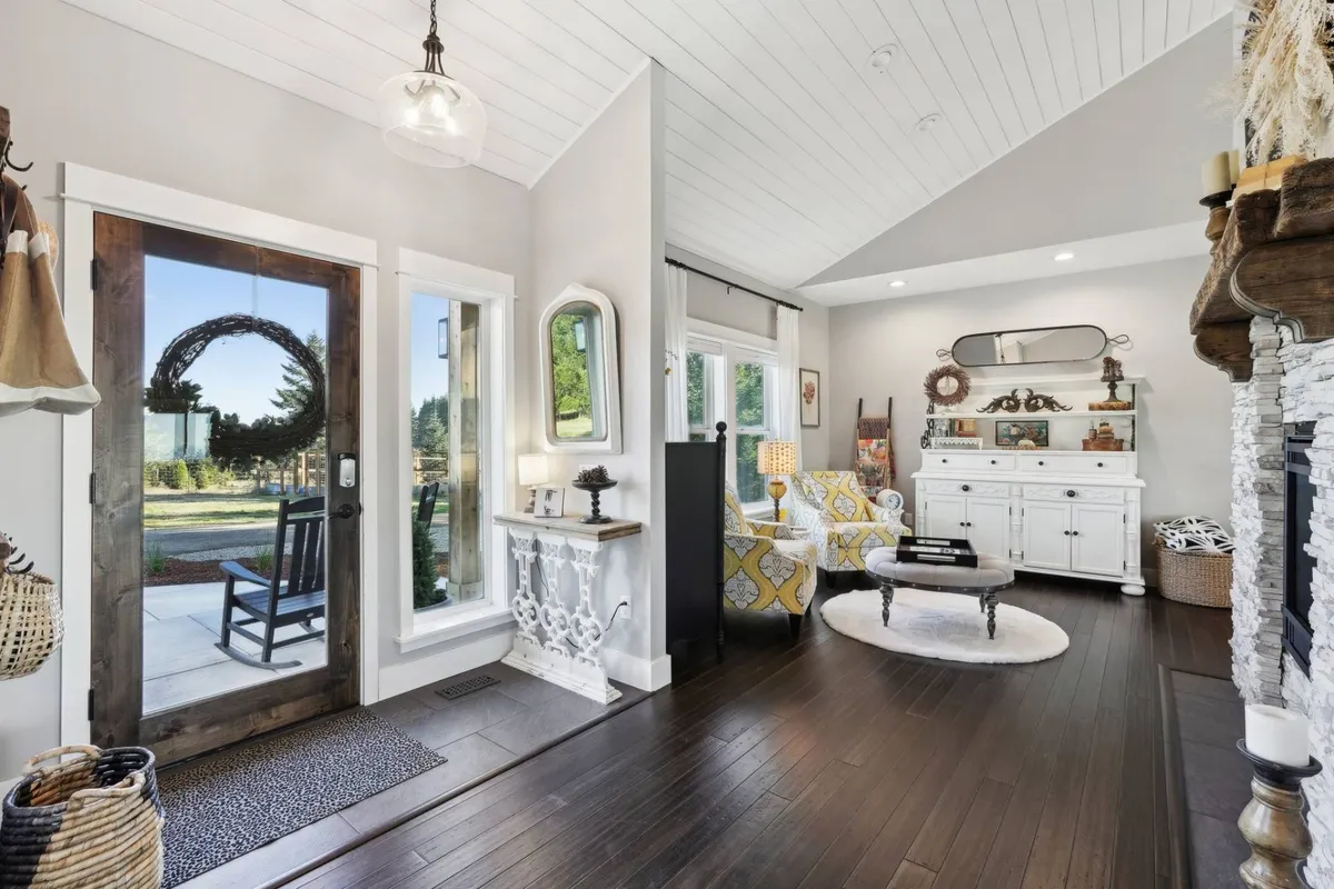 A cozy seating area just inside the front door, illuminated by natural light from large windows. The scene includes a fireplace, comfortable seating, and a decorative console table, with views to the outdoors visible through the glass.