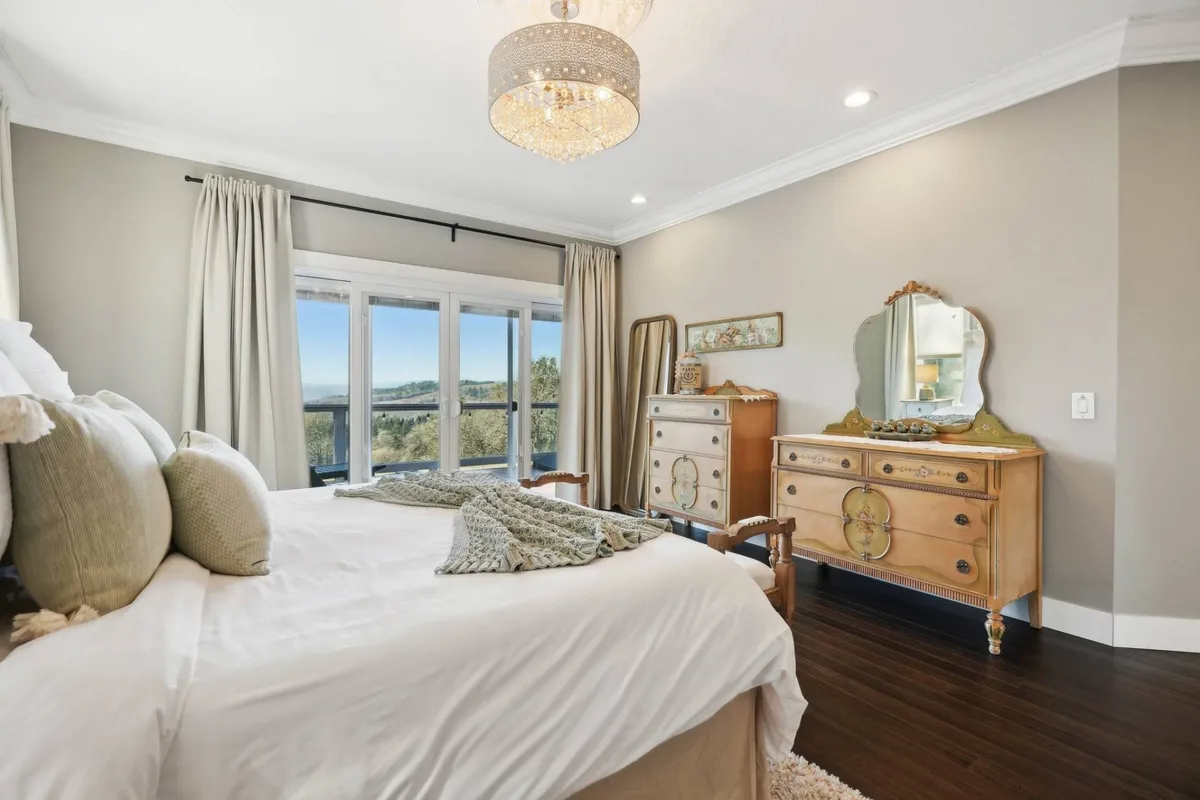 A serene bedroom featuring a plush bed with decorative pillows, elegant chandelier, large windows framing a beautiful view of the surrounding landscape, a wooden dresser, and a mirror. The soft color palette, natural light, and inviting textures create an atmosphere of relaxation and quiet retreat.