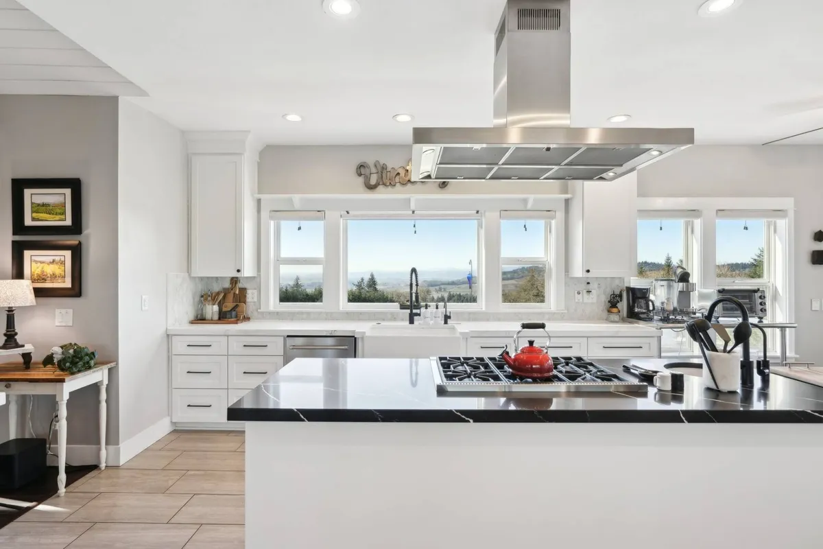 This image features a kitchen island with a sleek black countertop, high-end stainless steel appliances, and large windows that offer expansive views of the landscape. A decorative sign above the windows adds a touch of personality, while abundant natural light floods the space, creating a warm and inviting atmosphere ideal for cooking and gathering.