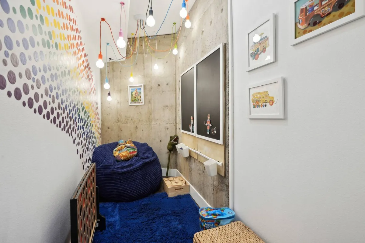 A playful, colorful room featuring a dotted accent wall, a cozy bean bag chair, whimsical hanging light bulbs, a blackboard, and framed artwork. The room is clearly designed for creative play, reading, and relaxation, with an inviting and imaginative atmosphere perfect for children or young-at-heart guests.
